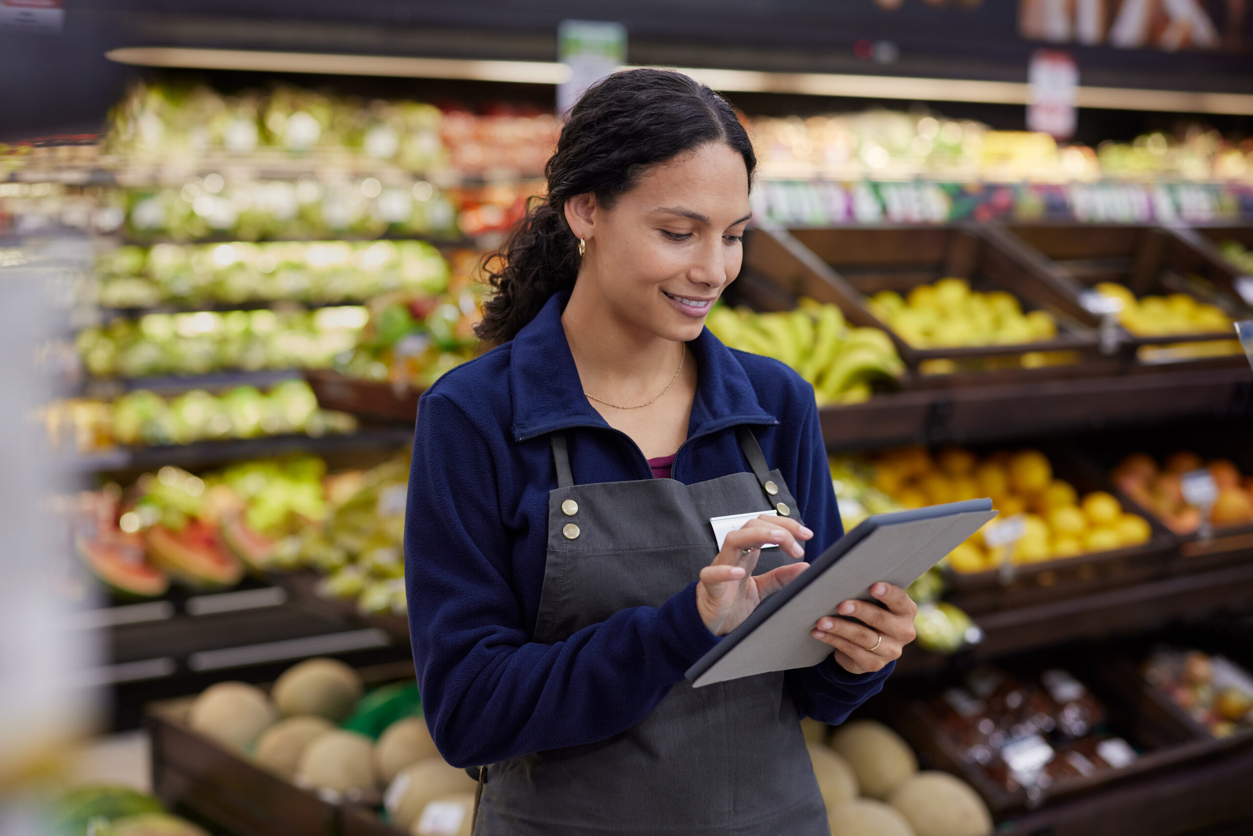 Store worker using tablet at supermarket Grocery store employee checking inventory on a digital tablet at supermarket. Young multiethnic woman in apron standing in front of fresh product shelves using touchscreen device. Smiling female worker using tablet while managing tasks in fruit section at grocery store.