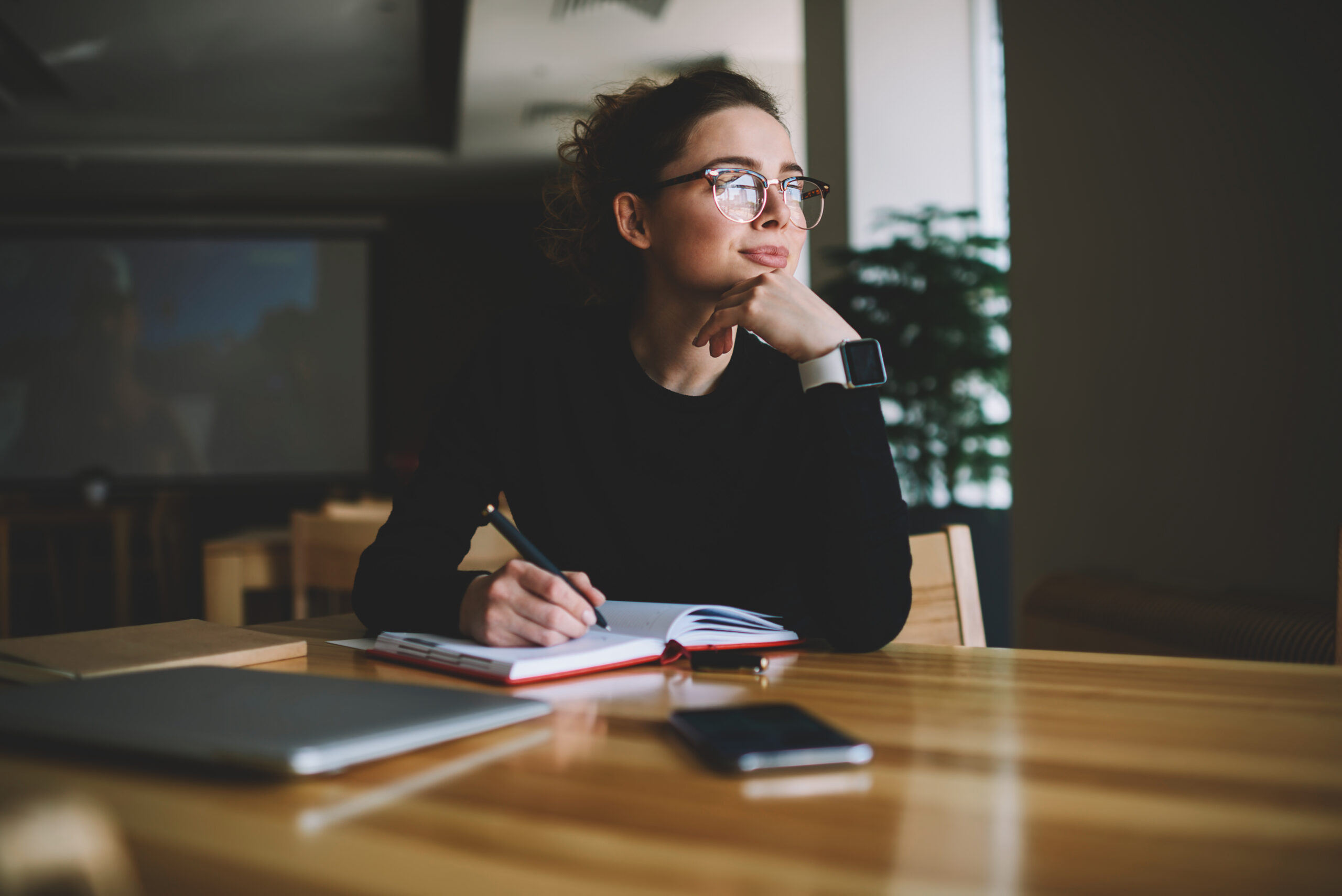 Thoughtful young author businesswoman and thinking on writing in Thoughtful young author businesswoman and thinking on writing interesting article.Woman in stylish eyeglasses with good lenses looking out of window and pondering new plans while noting them in notepad