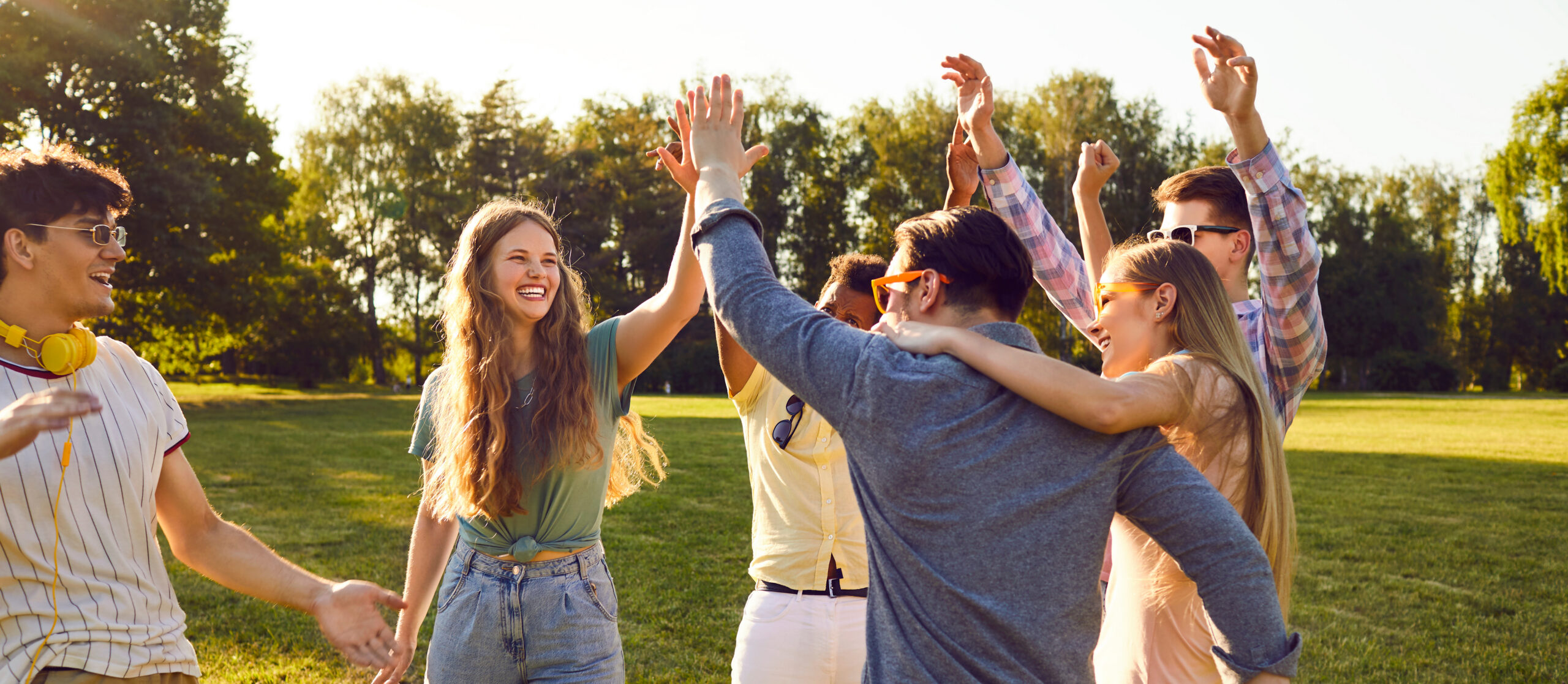 Group of happy cheerful young friends meeting in park and having fun all together Bunch of happy young friends all together having fun on warm sunny day in summer park. Diverse group of cheerful joyful positive people standing on green lawn, smiling and giving each other high five