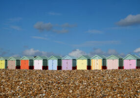 A row of ten colorful beach huts are in a line in the centre of the image in the foreground is a pebble beach above is a big blue sky with white clouds the sun is shining, Brighton, Sussex, United Kingdon, UK