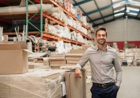 Portrait of a smiling warehouse manager leaning against some stock with piles of carpets stacked on shelves in the background
