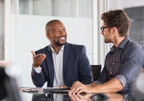 Two happy business colleagues at meeting in modern office interior. Successful african boss in a conversation with young employee in boardroom. Marketing team of two businessmen discussing strategy in meeting room.