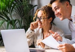 Portrait of colleagues working together in office with computer and plant in background. Smiling woman and young boy work. Small business family lifestyle. Happy team worker two people using laptop